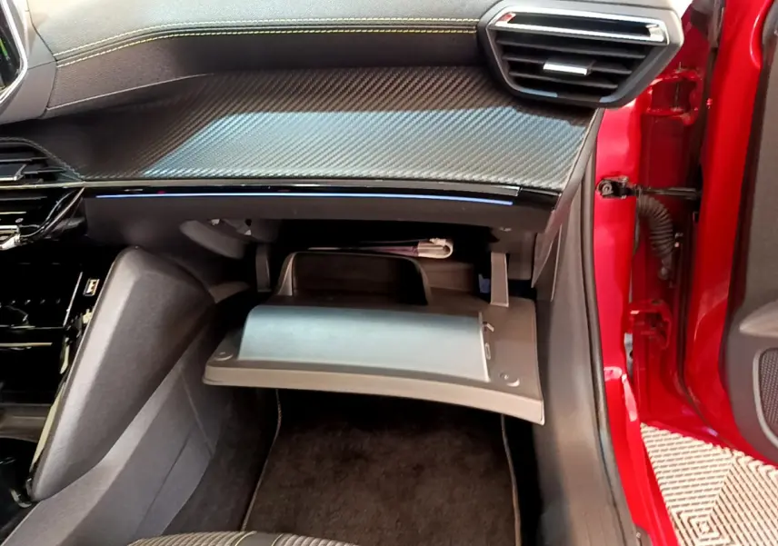 Vue intérieure côté passager d'une Peugeot 208 rouge 2020 avec boîte à gants ouverte et tableau de bord noir à insert carbone.
