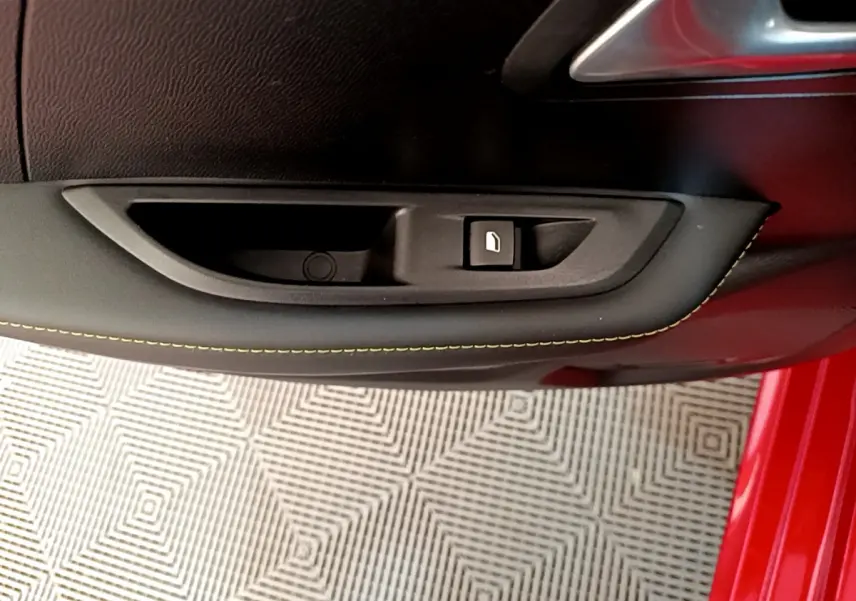 Gros plan sur la poignée intérieure côté gauche d'une Peugeot 208 rouge avec couture jaune visible sur la portière.