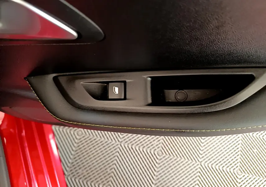 Gros plan sur la commande de lève-vitre côté gauche dans la porte intérieure noire d'une Peugeot 208 rouge.