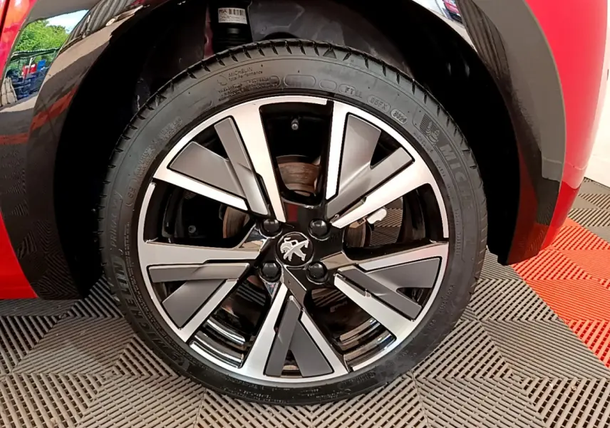 Gros plan sur la roue avant droite de la Peugeot 208 rouge, mettant en valeur la jante alliage au design moderne.