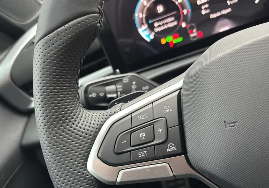 Gros plan sur le volant cuir perforé multifonctions du Volkswagen T-Roc gris loup, avec tableau de bord numérique en arrière-plan flou.