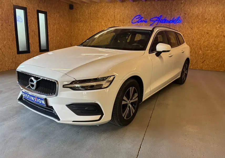 Volvo V60 D3 blanche vue 3/4 avant droit dans un showroom avec mur en bois et enseigne lumineuse bleue.