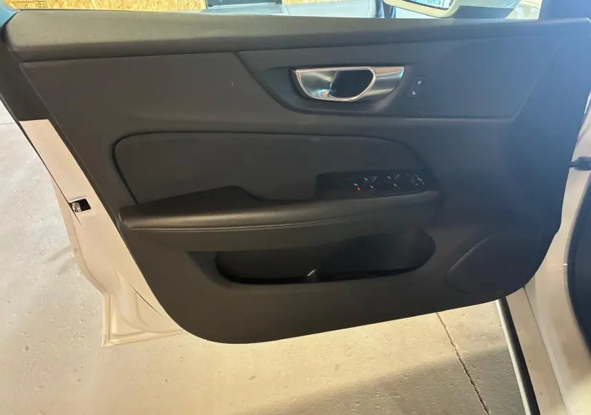 Vue intérieure de la porte avant gauche noire d’une Volvo V60 blanche, avec commandes de vitres et poignée métal.