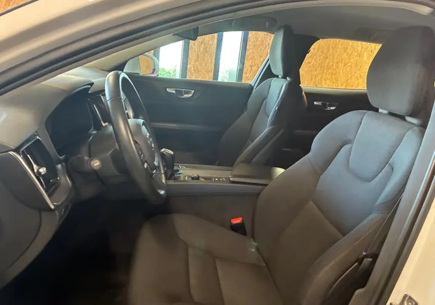 Vue intérieure côté conducteur du Volvo V60 blanc, mettant en valeur les sièges en tissu noir et le volant multifonction.