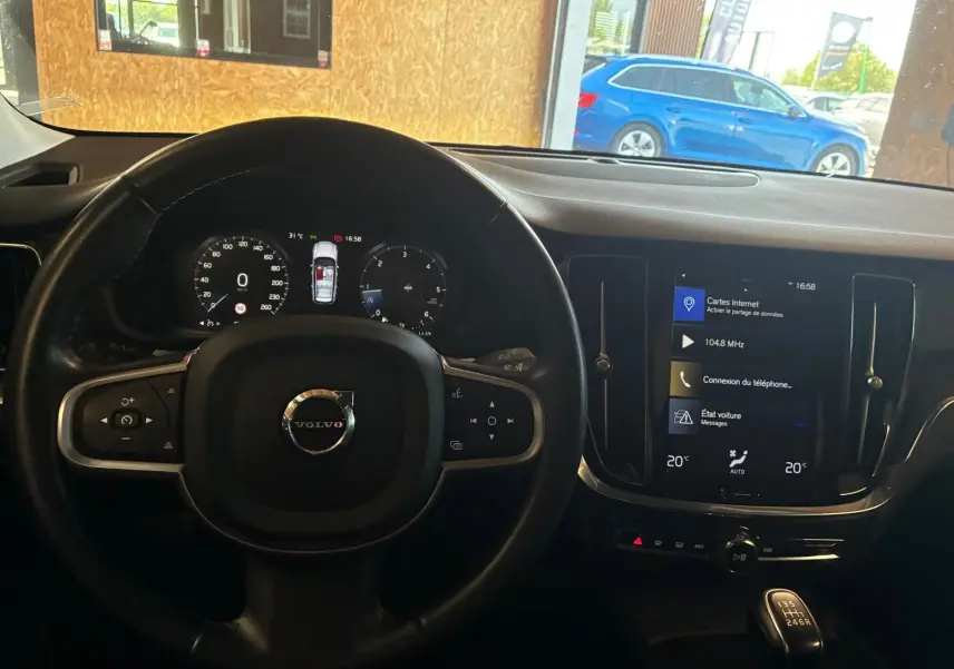 Vue intérieure centrée sur le volant et le tableau de bord noir d'une Volvo V60 blanche, avec écran tactile allumé.