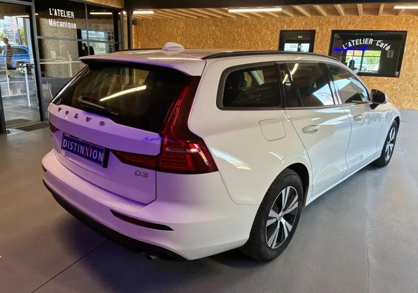 Vue 3/4 arrière droite d’un break Volvo V60 D3 blanc 2020 avec feux arrière LED et toit ouvrant en intérieur d’atelier.