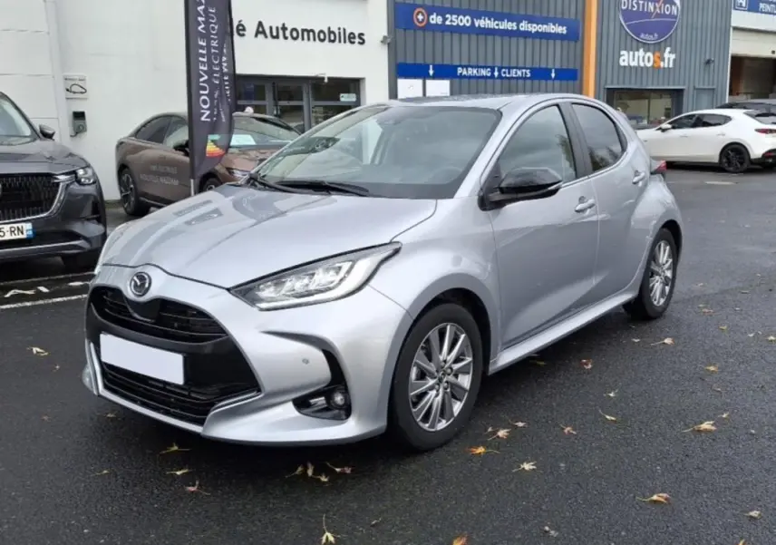 Vue 3/4 avant d'une Mazda2 Hybrid argent 2022 garée devant un concessionnaire, avec calandre noire et phares LED.