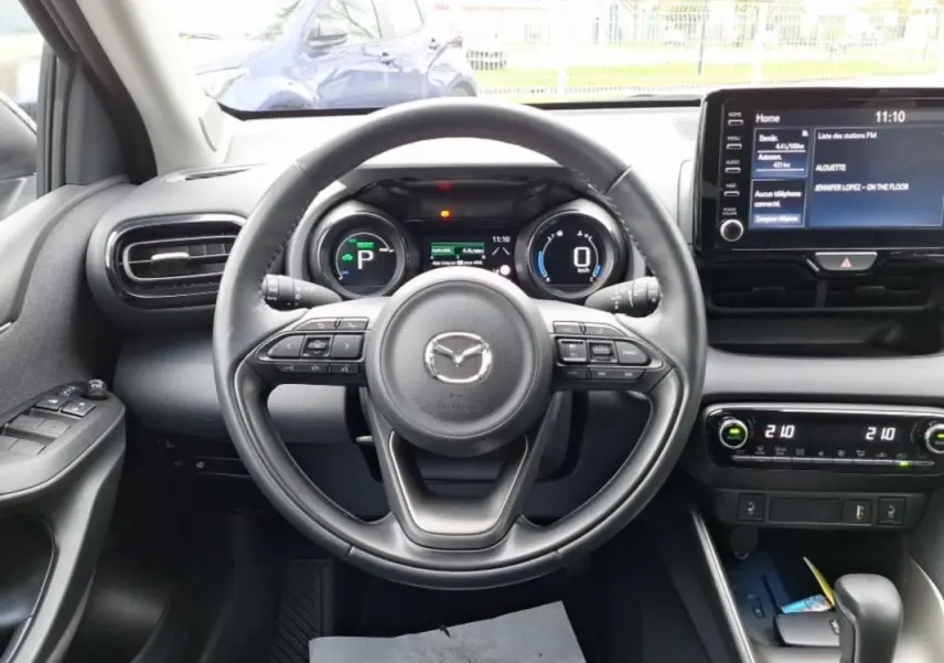 Vue intérieure centrée sur le volant de la Mazda2 hybride 2022, avec tableau de bord numérique et écran tactile visible.