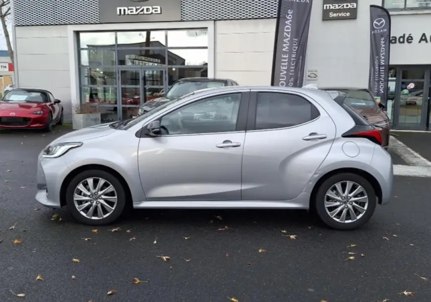 Profil côté gauche d'une Mazda2 hybride 2022 argent garée devant un concessionnaire Mazda avec jantes alliage.