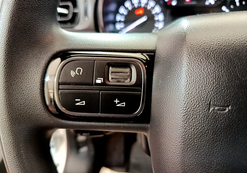 Gros plan sur les commandes de volume et téléphone au volant noir de la Citroën C3 blanche, tableau de bord flou en arrière-plan.