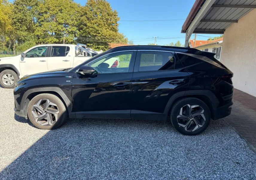 Vue de profil côté gauche d’un Hyundai Tucson noir 2022 garé sur gravier, avec barres de toit et jantes distinctives.