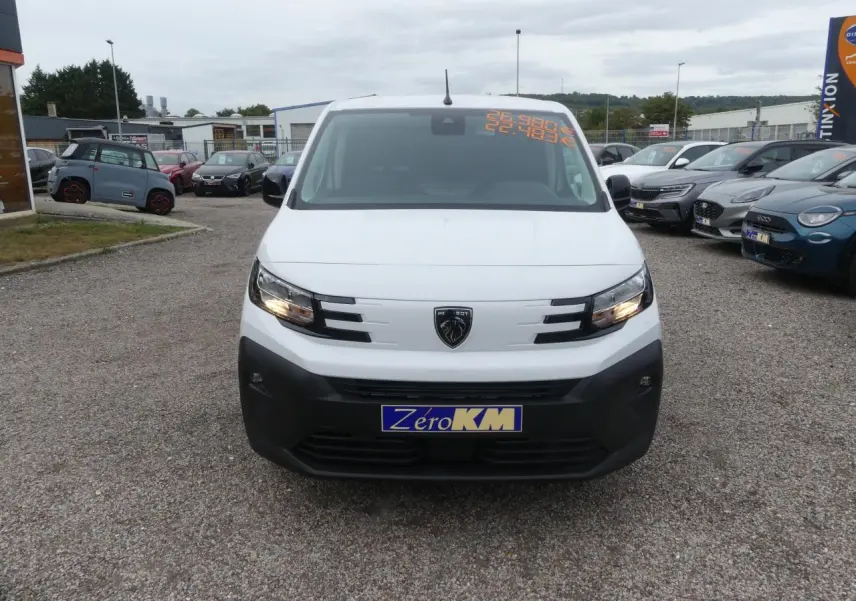 Vue de face d'un Peugeot Partner blanc avec pare-chocs noir et plaque d'immatriculation Zéro KM sur un parking.