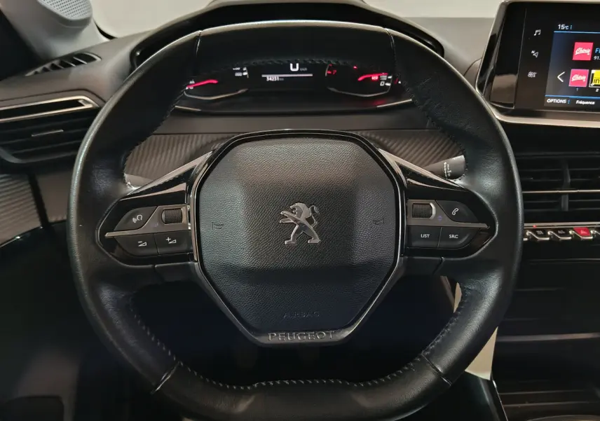 Vue rapprochée du volant cuir noir de la Peugeot 208 PureTech 100, avec tableau de bord digital et écran tactile visible.