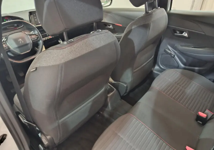 Vue intérieure arrière de la Peugeot 208 blanche, montrant les sièges noirs avec surpiqûres rouges et le tableau de bord avant.