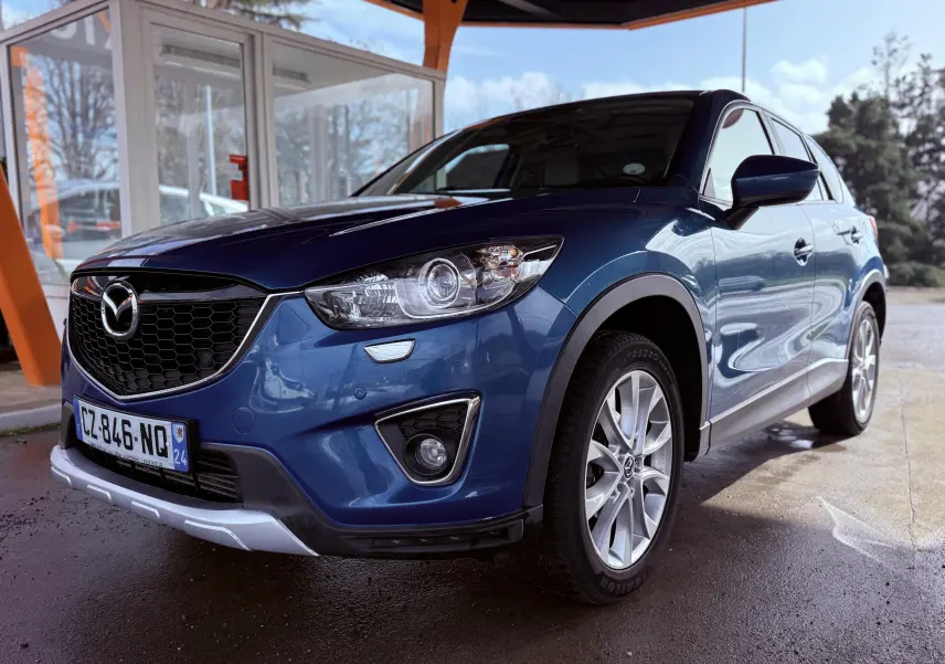 Vue 3/4 avant droit d'un Mazda CX-5 bleu 2013 avec calandre noire et jantes argentées, sous un abri extérieur.