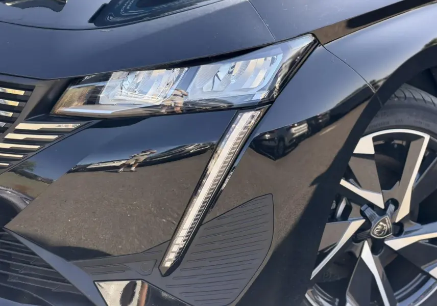 Gros plan sur l'avant droit noir Perla de la Peugeot 308 Hybrid 2025, mettant en valeur le phare LED et la jante alliage 17'' Halong.