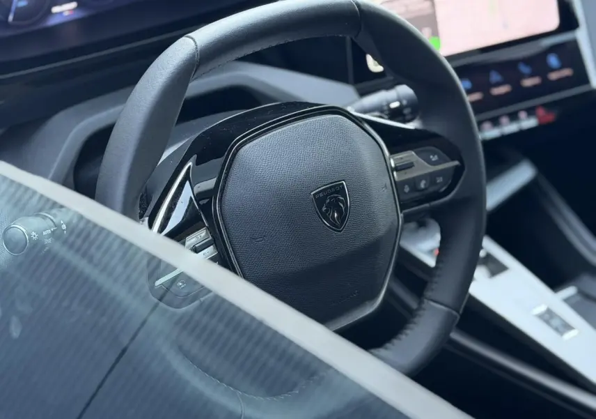 Gros plan sur le volant cuir noir de la Peugeot 308 Hybrid 2025 avec tableau de bord et écran tactile en arrière-plan.