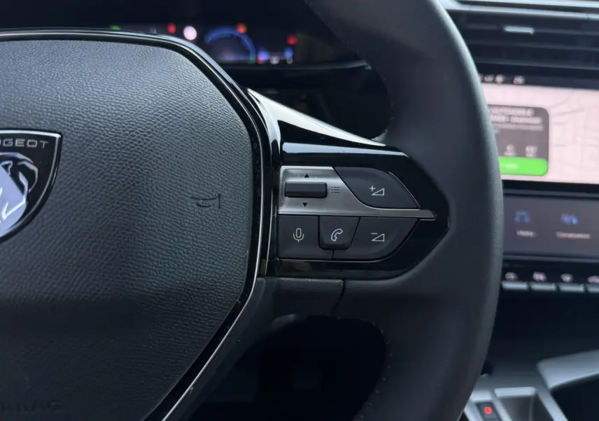 Gros plan sur le volant noir de la Peugeot 308 Hybrid 2025, avec commandes intégrées et écran tactile de navigation en arrière-plan.