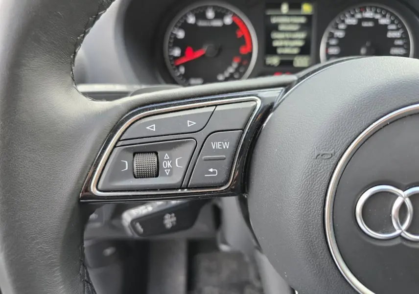 Gros plan sur la partie gauche du volant cuir noir de l'Audi Q2 2023 avec commandes multifonctions et tableau de bord flou.