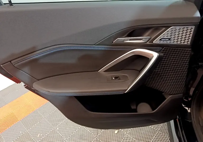 Gros plan sur la porte côté gauche du BMW X1 noir, avec surpiqûres bleues et haut-parleur Harman Kardon intégré.