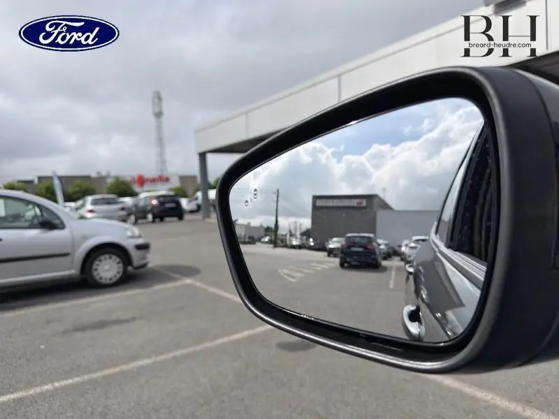 Rétroviseur côté droit d’un Ford Kuga gris Magnetic Fashion, vue partielle avec reflet du parking et ciel nuageux.