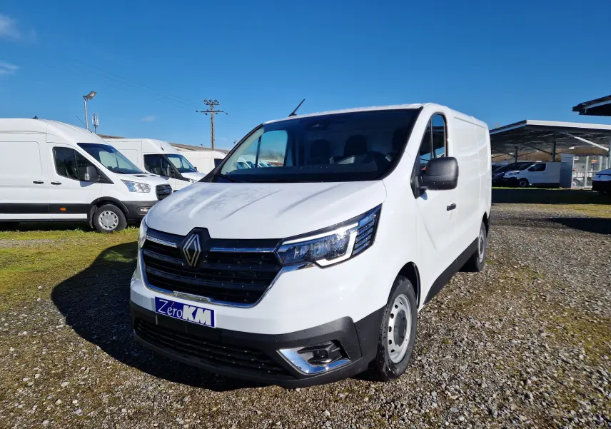 Renault Trafic Fourgon blanc en 3/4 avant droit sur parking extérieur, avec calandre noire et phares LED visibles.