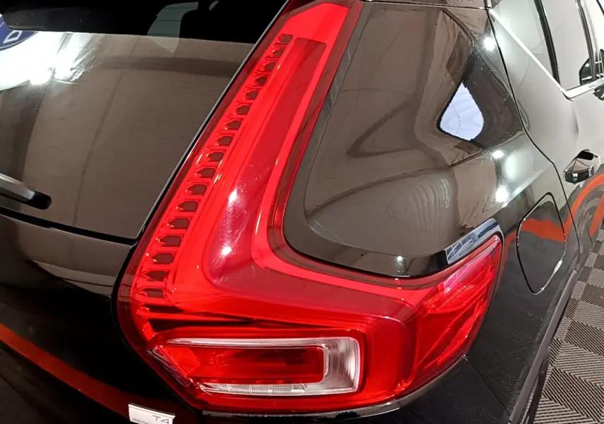 Gros plan sur le feu arrière droit rouge vif d’un Volvo XC40 Recharge T4 noir brillant, avec reflet du plafond visible.