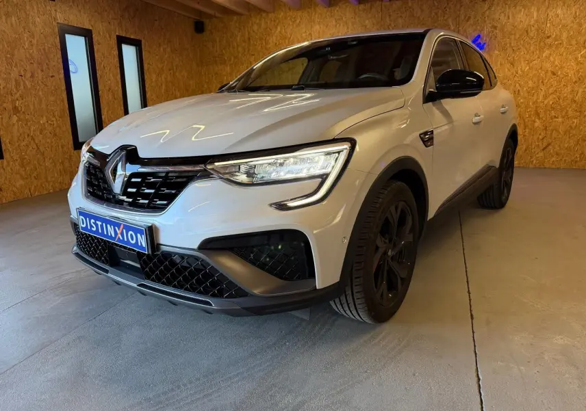 Vue avant d'un Renault Arkana blanc nacré avec calandre noire et phares LED allumés en intérieur showroom.