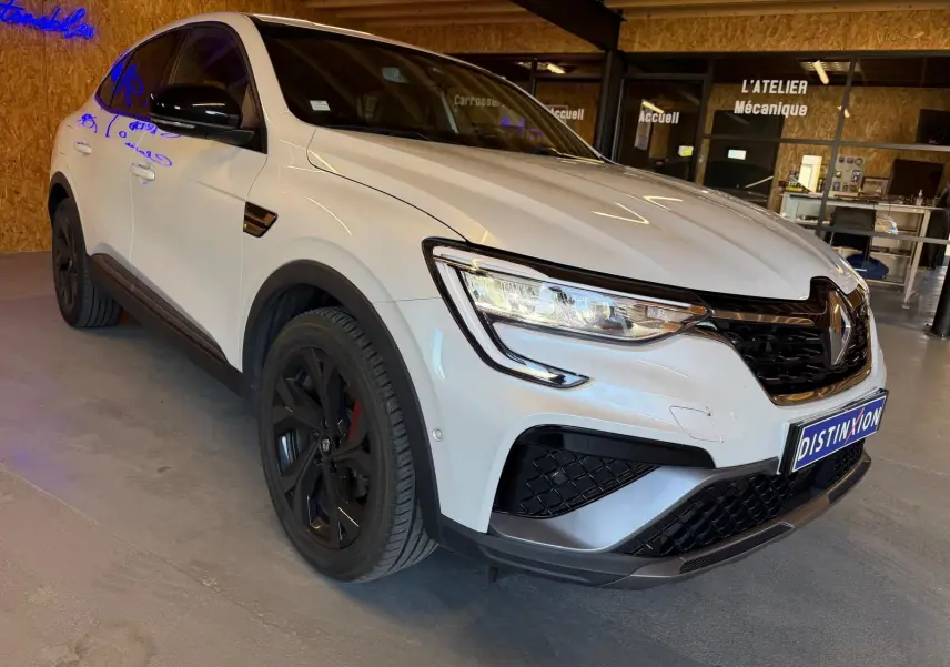 Renault Arkana blanc nacré en 3/4 avant droit, avec calandre noire et jantes alu noires, stationné en intérieur.