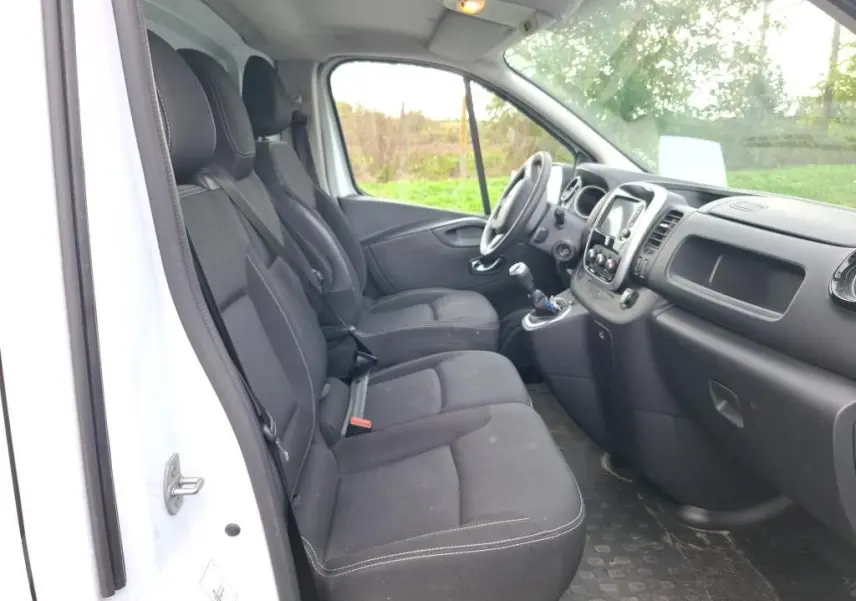 Vue intérieure côté conducteur du Renault Trafic Fourgon blanc, montrant les sièges noirs et le tableau de bord moderne.