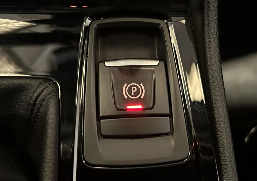 Gros plan sur le bouton de frein à main électronique avec témoin rouge dans l’habitacle d’une BMW Série 2 Active Tourer blanche.