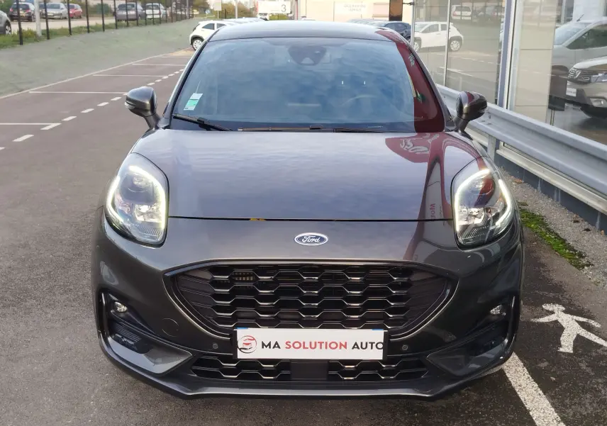 Vue frontale d'un Ford Puma gris métallisé avec calandre noire et phares allumés sur un parking extérieur.