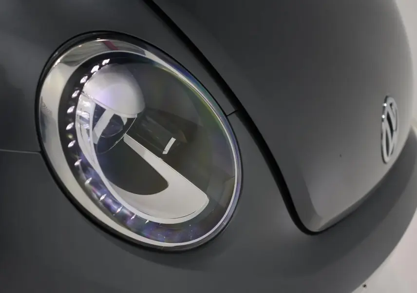 Gros plan sur le phare avant gauche gris d’une Volkswagen Coccinelle 2016 avec logo VW visible sur le capot.