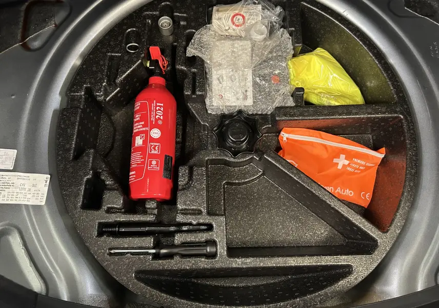 Vue plongeante sur le coffre de la Volkswagen Coccinelle gris, montrant le rangement avec extincteur rouge et trousse de secours orange.
