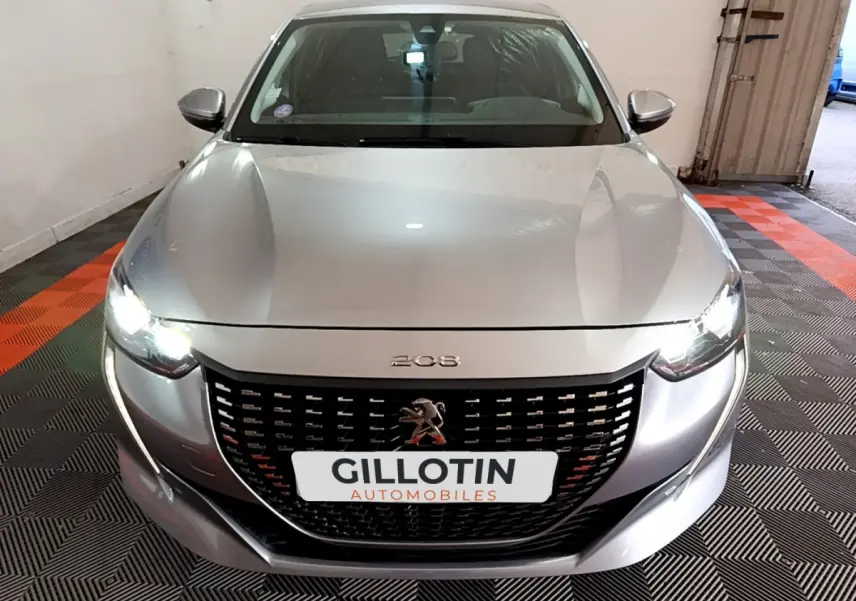 Vue avant d'une Peugeot 208 gris clair avec phares LED allumés dans un garage fermé.