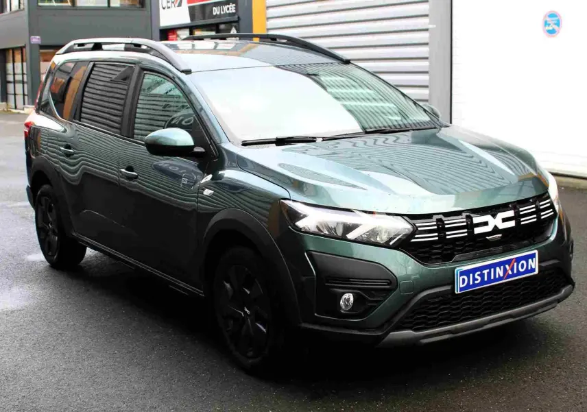 Vue 3/4 avant d'un Dacia Jogger vert cèdre métallisé avec calandre noire et jantes foncées, stationné devant un bâtiment.