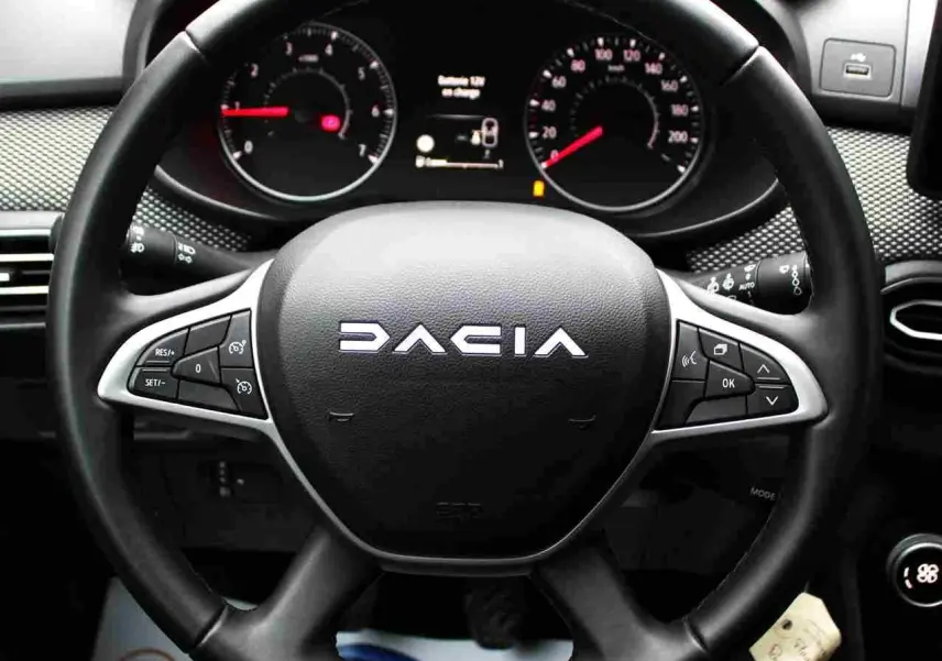 Vue rapprochée du volant noir DACIA avec commandes intégrées, tableau de bord et affichage numérique en arrière-plan.