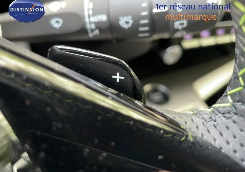 Gros plan sur la palette de changement de vitesse droite en plastique noir et volant cuir gris avec surpiqûres vertes.
