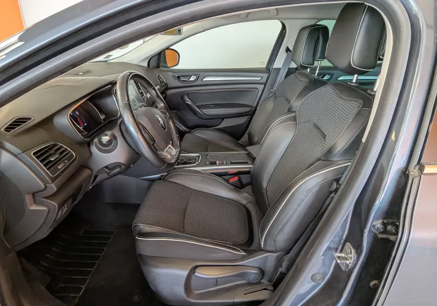 Intérieur de la Renault Mégane IV gris, vue côté conducteur, sièges noirs avec surpiqûres blanches et tableau de bord moderne.