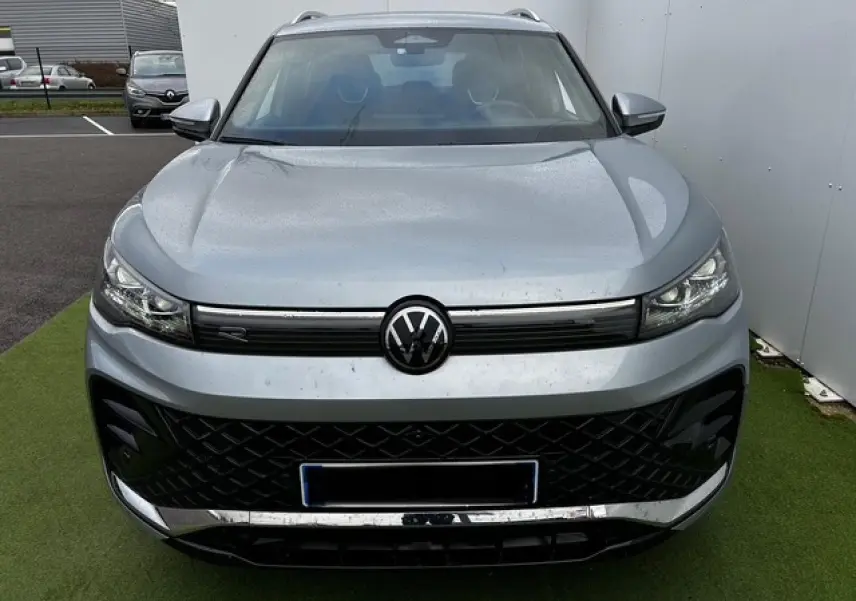 Vue frontale d'un Volkswagen Tiguan III gris clair avec calandre noire et logo VW central sur fond urbain.
