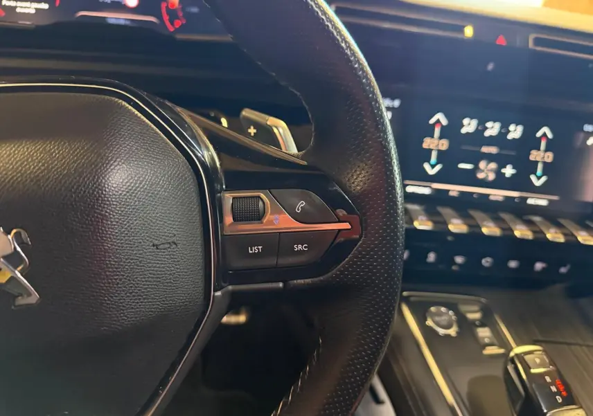 Gros plan sur le volant cuir noir et les commandes multifonctions, avec écran tactile et console bois gris en arrière-plan.