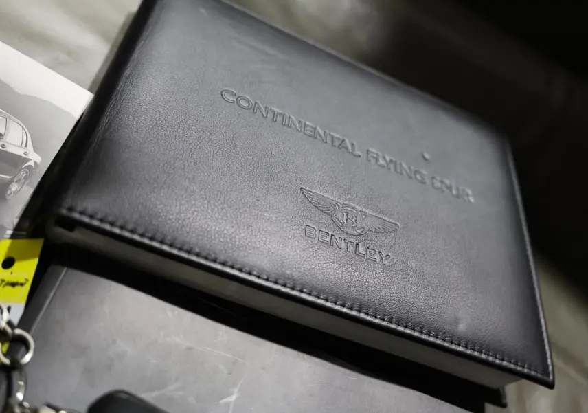 Image montrant un carnet d'entretien en cuir noir embossé Bentley Continental Flying Spur, posé sur une surface grise.