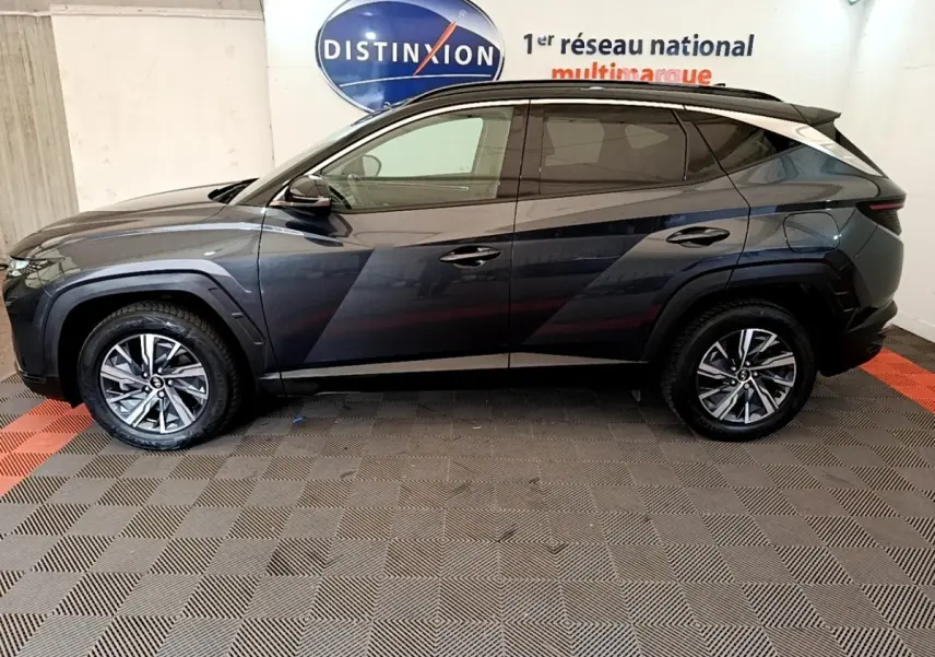 Vue de profil côté gauche d'un Hyundai Tucson 1.6 Hybrid 230 Creative gris foncé avec jantes alliage en intérieur showroom.