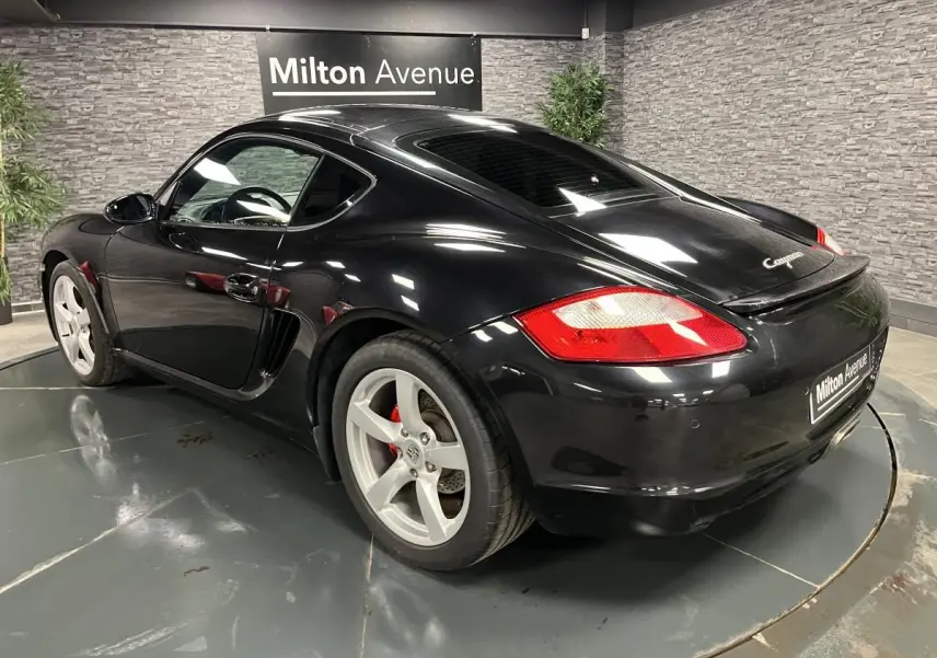 Vue 3/4 arrière droite d'une Porsche Cayman noire avec jantes argentées et feux arrière rouges, en intérieur showroom.
