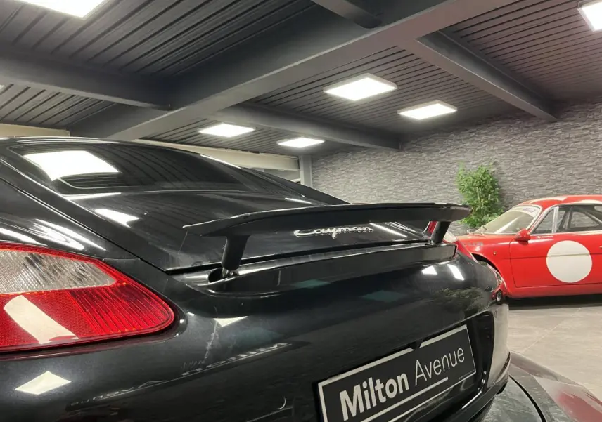 Vue arrière droite d'une Porsche Cayman noire avec aileron arrière déployé dans un showroom intérieur.