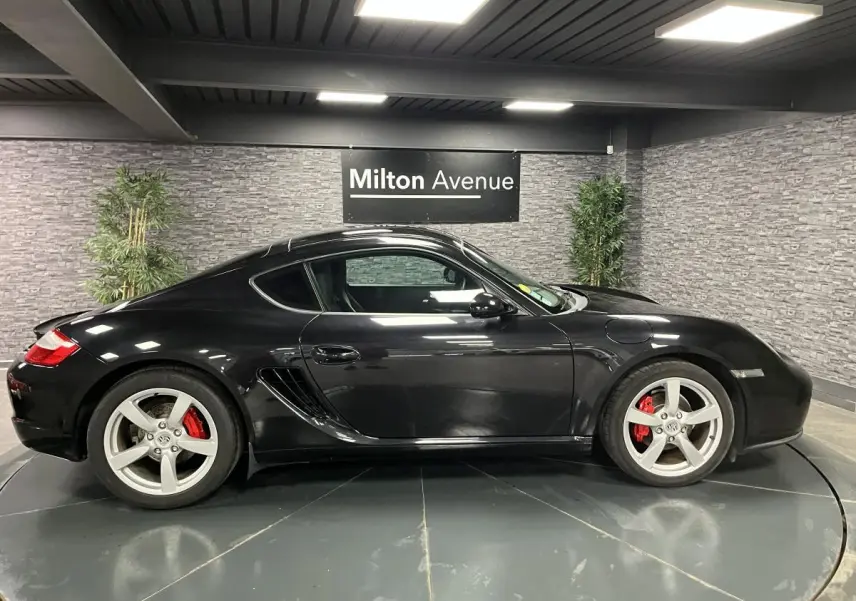 Profil côté gauche d'une Porsche Cayman noire 2008 avec jantes argentées et étriers de frein rouges en intérieur showroom.
