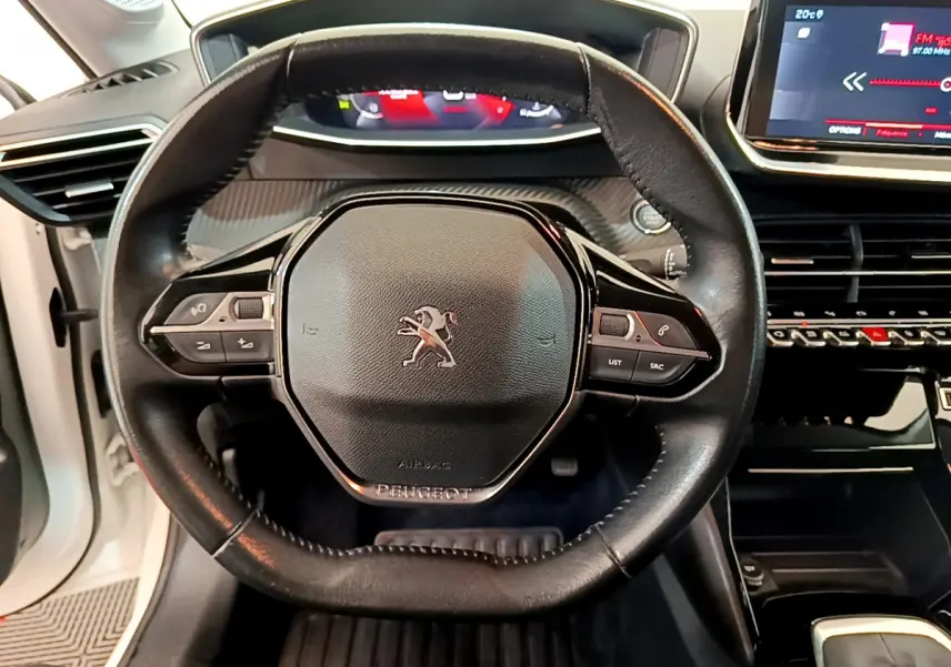 Vue rapprochée du volant cuir noir de la Peugeot 208 blanche, avec tableau de bord digital et écran tactile central visible.
