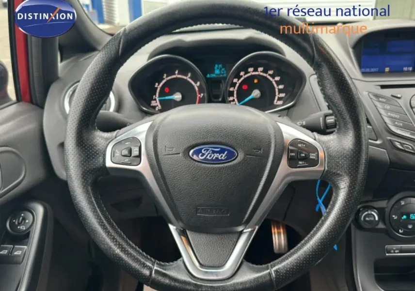 Vue centrée sur le volant noir perforé de la Ford Fiesta rouge, avec tableau de bord et commandes visibles.