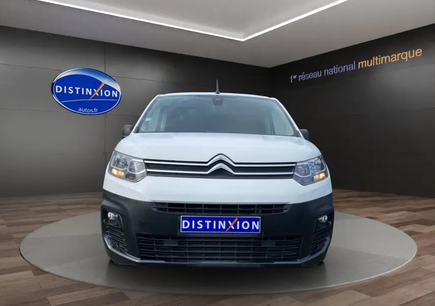 Vue de face d'un Citroën Berlingo blanc avec pare-chocs noir et feux allumés en showroom.