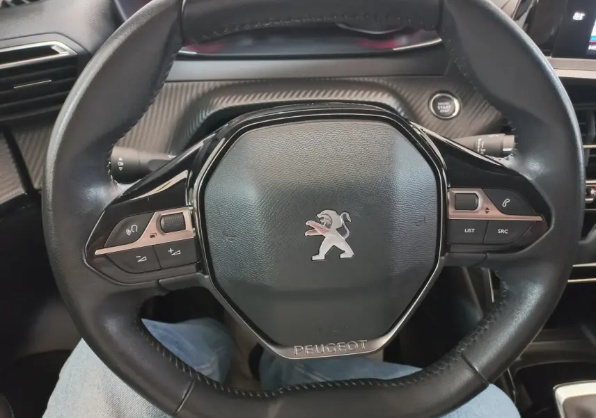 Vue rapprochée du volant cuir noir de la Peugeot 208 PureTech 100 S&S Allure avec commandes intégrées et bouton démarrage visible.